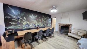 Un espace repas comprenant une longue table en bois, six chaises, une fresque représentant des branches d’olivier, une cheminée et un canapé.