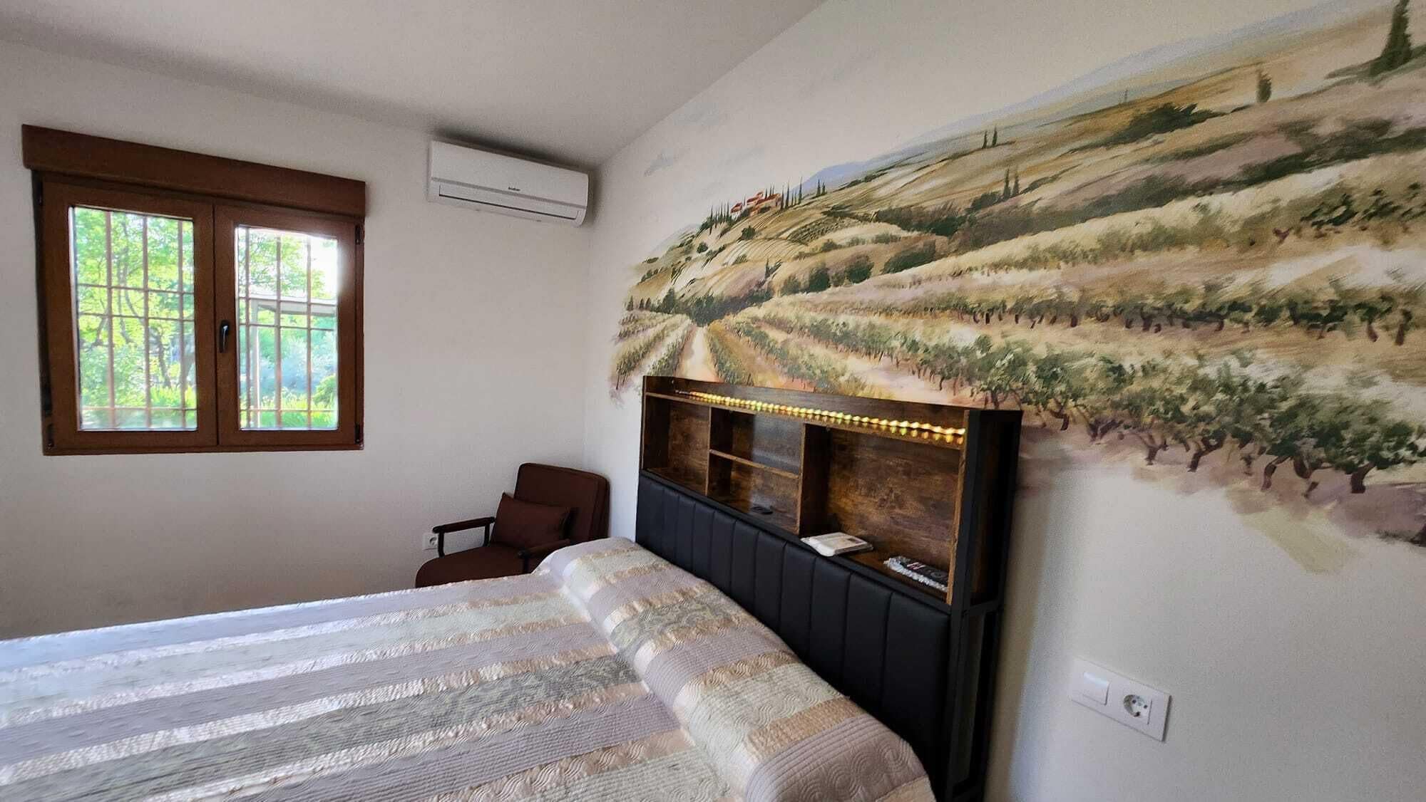 Ein Schlafzimmer mit einem großen Bett, einem Holzkopfende, einem Fenster und einem Wandgemälde von einem Weinberg.