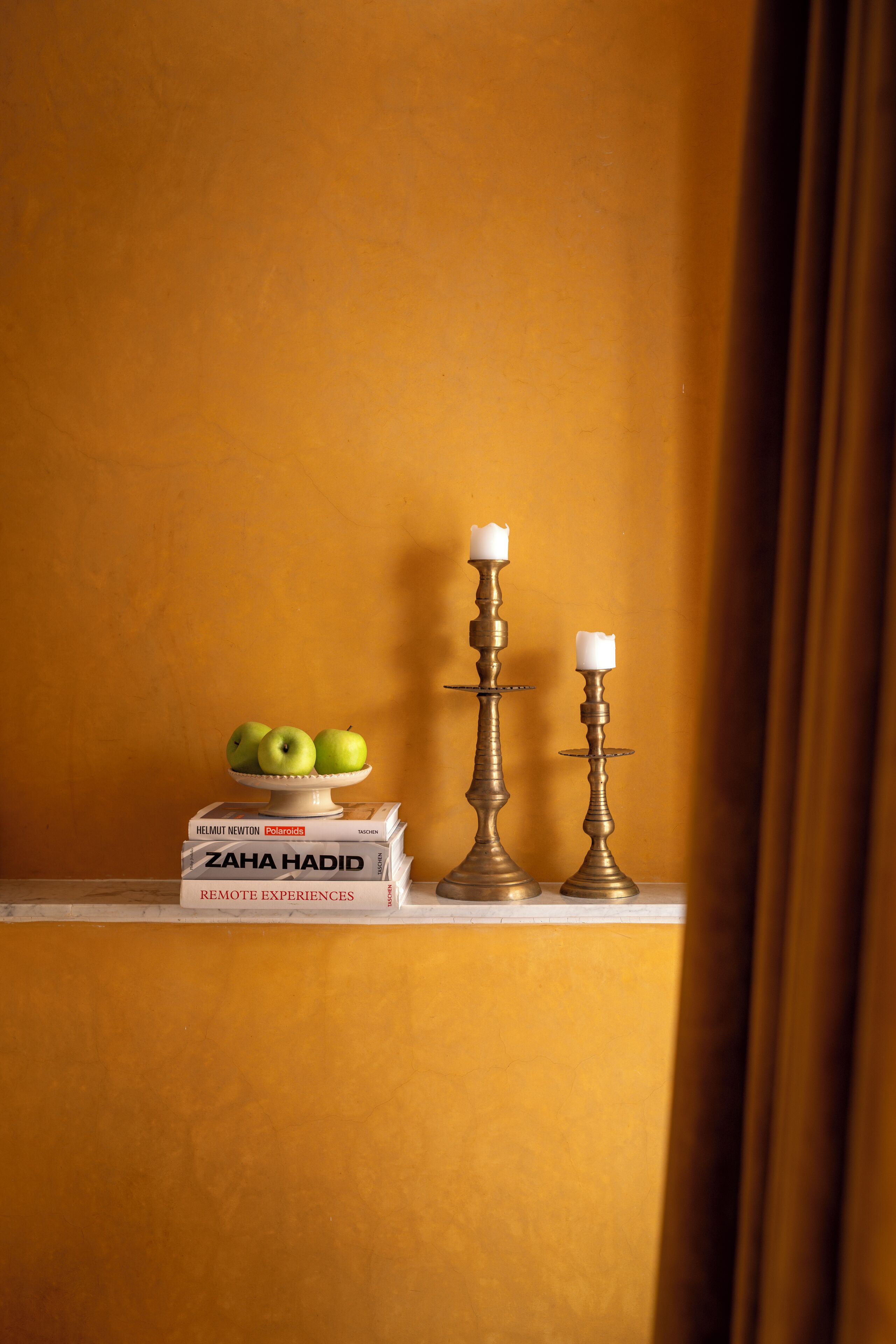 A shelf with books, apples, and two candle holders against a yellow wall.