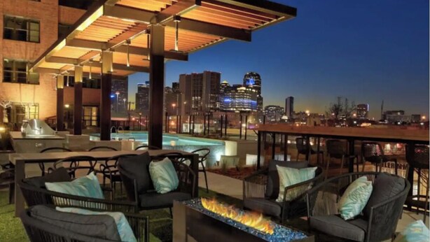 An outdoor seating area with a fire feature, patio furniture, and a view of the city skyline at dusk. - Clover Street Lofts (Dallas)