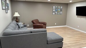Ein Wohnzimmer mit einem grauen Ecksofa, einem braunen Ledersessel, einer Lampe und einer Wand mit gerahmten Bildern.