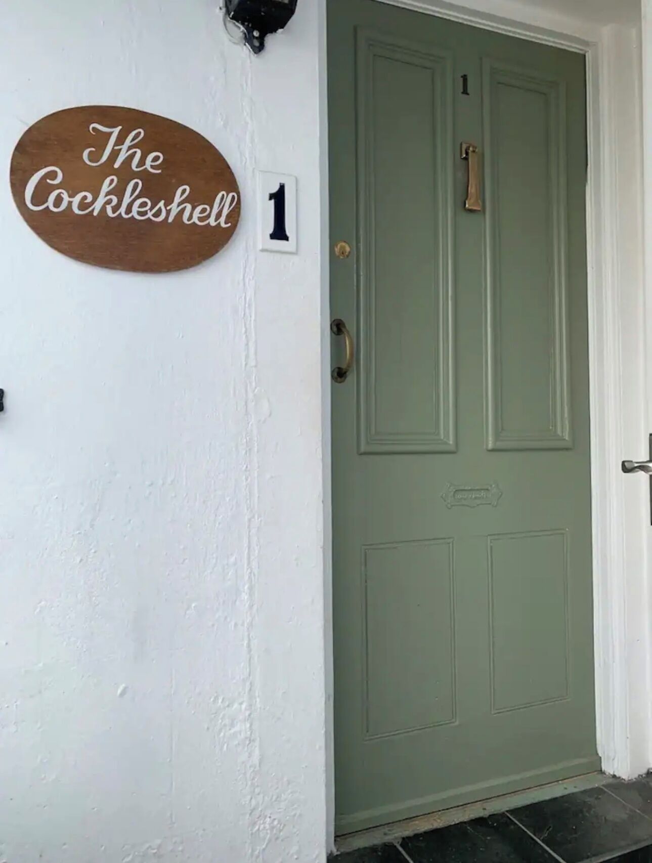 A green door with a brass handle and a brass knocker, a white wall with a sign reading "The Cockleshell 1", and a small number "1" above the door.