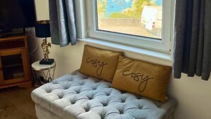 A room with a grey tufted sofa, two cushions with the word "cosy," a small side table with a lamp, a framed picture on the wall, and a window with a view of a marina.