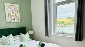 Una cama bien tendida con ropa de cama verde y dos toallas verdes, un cabecero verde, una ventana con vistas a barcos y vegetación, y una pared con una obra de arte con diseño de corazón.