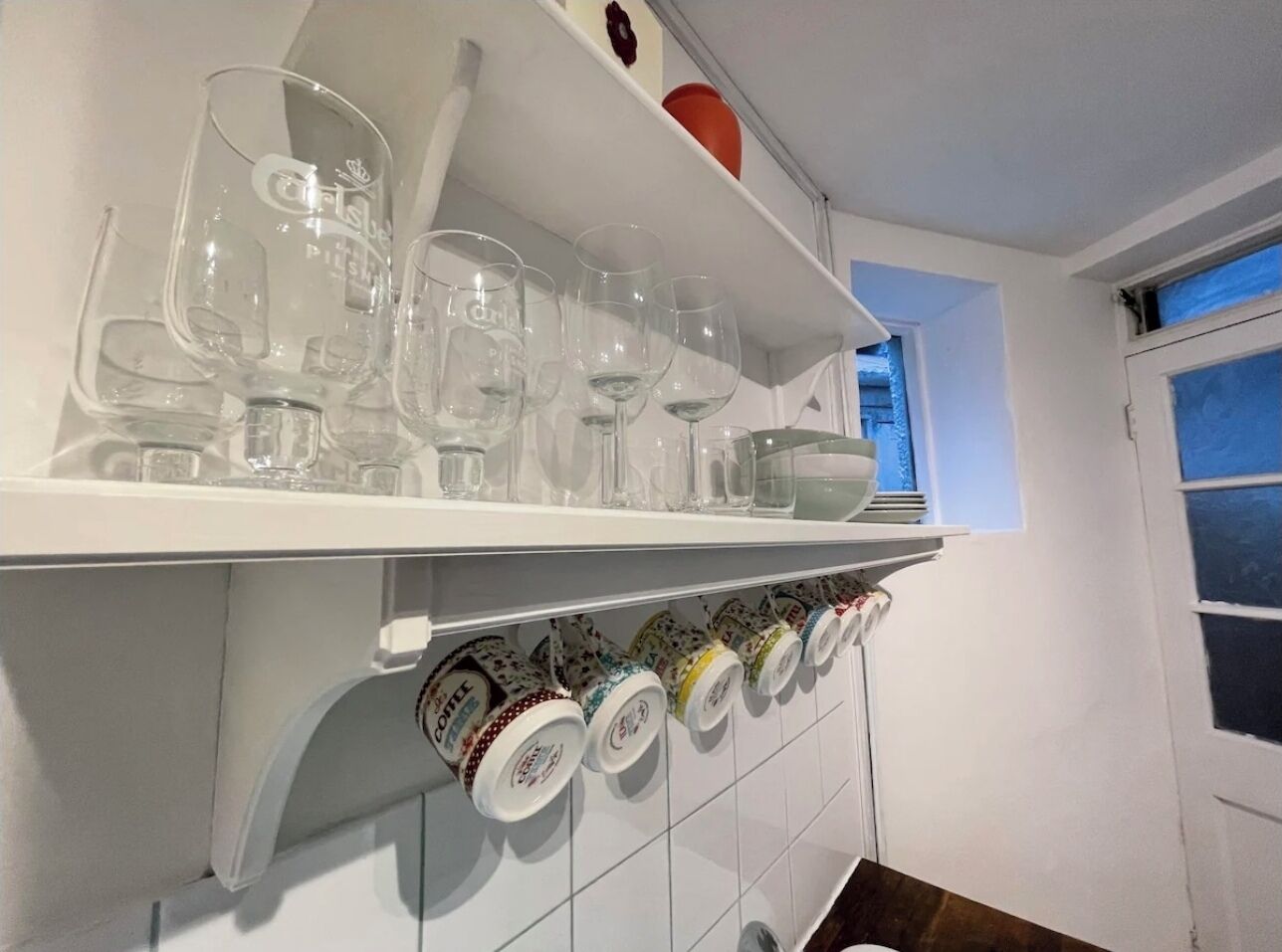 A kitchen with a white shelf holding wine glasses and a row of coffee mugs below.