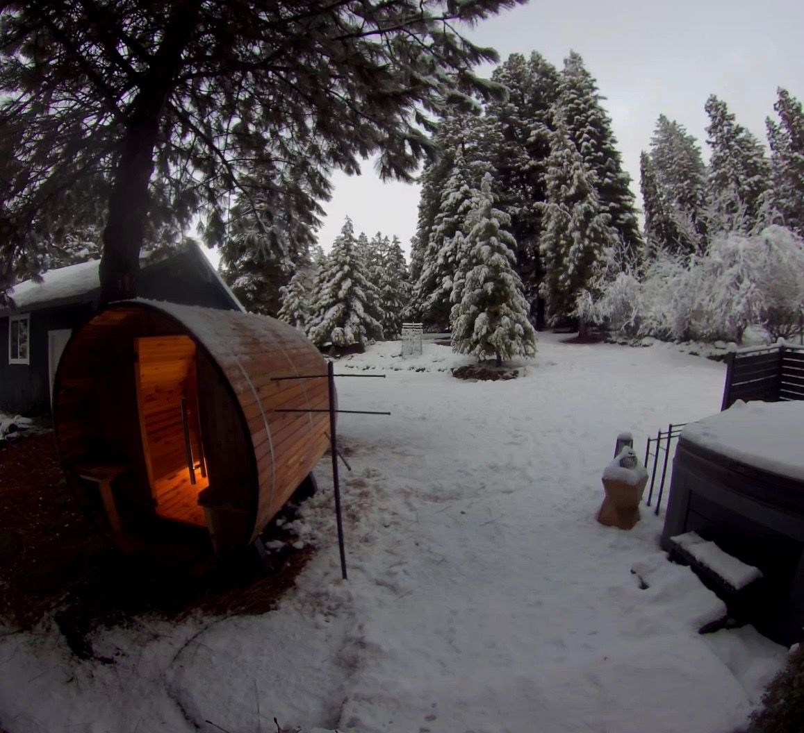 A snow-covered outdoor area with a wooden structure, possibly a sauna or hot tub, surrounded by snow-laden trees.