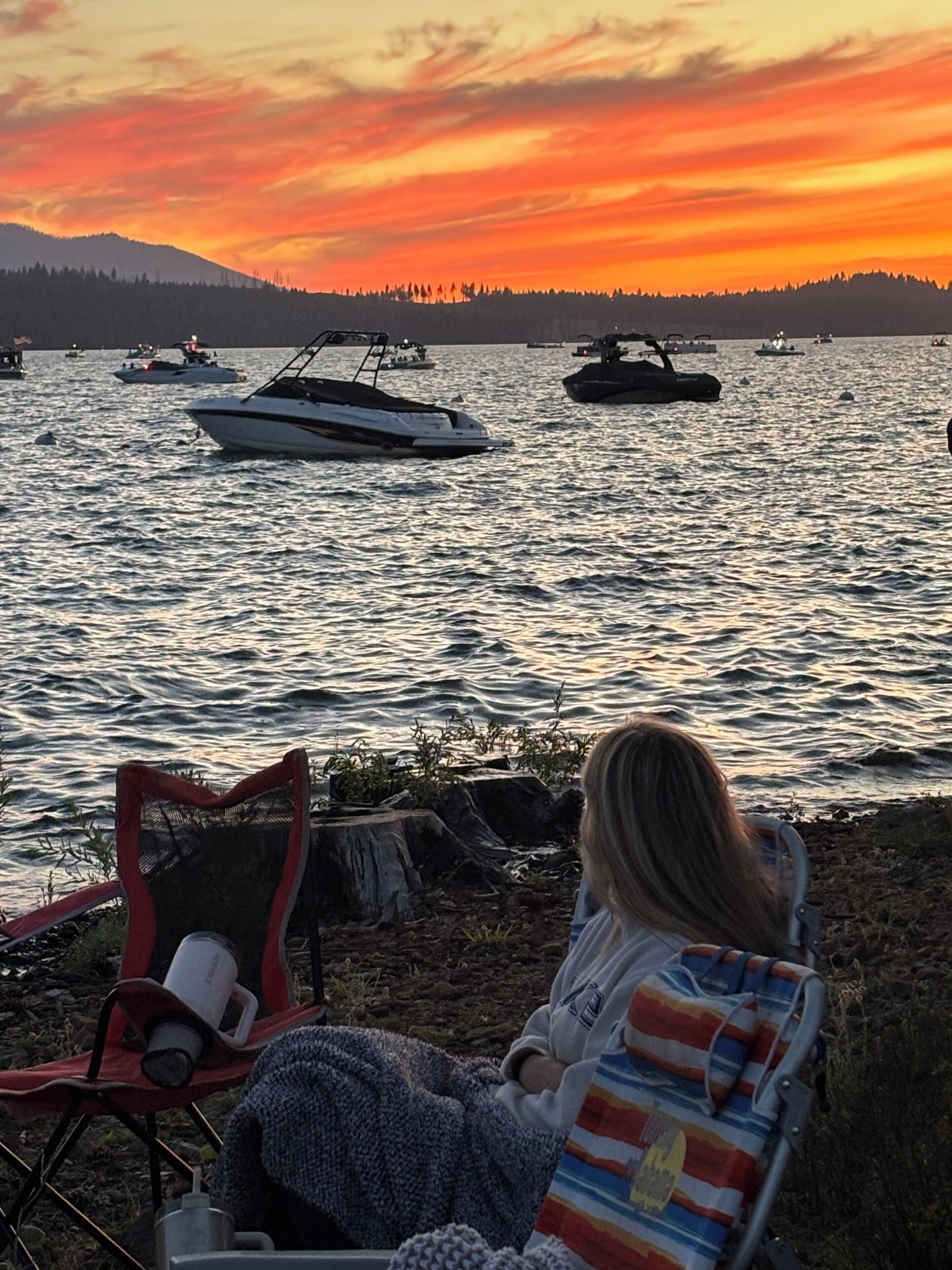A person seated on a chair by a lakeside during sunset, with boats on the water and a forested hill in the background.