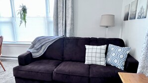 Een donkere bank met een deken en gekleurde kussens, een houten bijzettafeltje, een lamp en een raam met jaloezieën en een hangplant.
