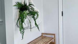 Een witte muur met een zwarte kapstok waarop een witte doek en groene planten hangen. Een houten bankje staat ervoor.