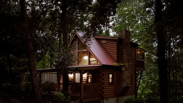 Une cabane en rondins dotée d’un balcon et d’un intérieur éclairé, entourée d’arbres.