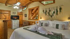 Une chambre avec un grand lit, des poutres apparentes en bois au plafond, un ventilateur de plafond, une cheminée et une décoration murale en macramé.