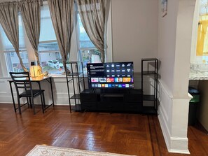 A living room with a flat-screen TV on a stand, a wooden dining table with chairs, and a window with curtains.
