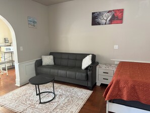 Sala de estar com sofá cinzento, mesa redonda pequena, tapete estampado, mesa de cabeceira branca com gavetas e cama com cobertor vermelho.