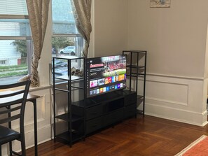 Un televisor sobre un soporte negro, con la pantalla mostrando varios canales, ubicado en una habitación con suelo de madera y una ventana con cortinas.