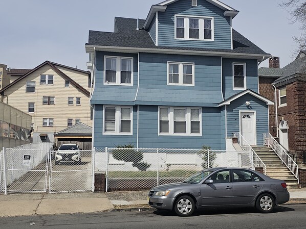 Uma casa de dois andares com fachada azul e detalhes brancos. Um carro está estacionado em frente, e uma cerca com uma placa de 'Proibido Entrar' é visível.