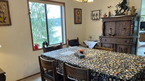A dining area with a floral tablecloth, wooden chairs, a chandelier, and a view through a window. - Spacious river facing 4-bedroom house in charming Waldport with WiFi. (Waldport)