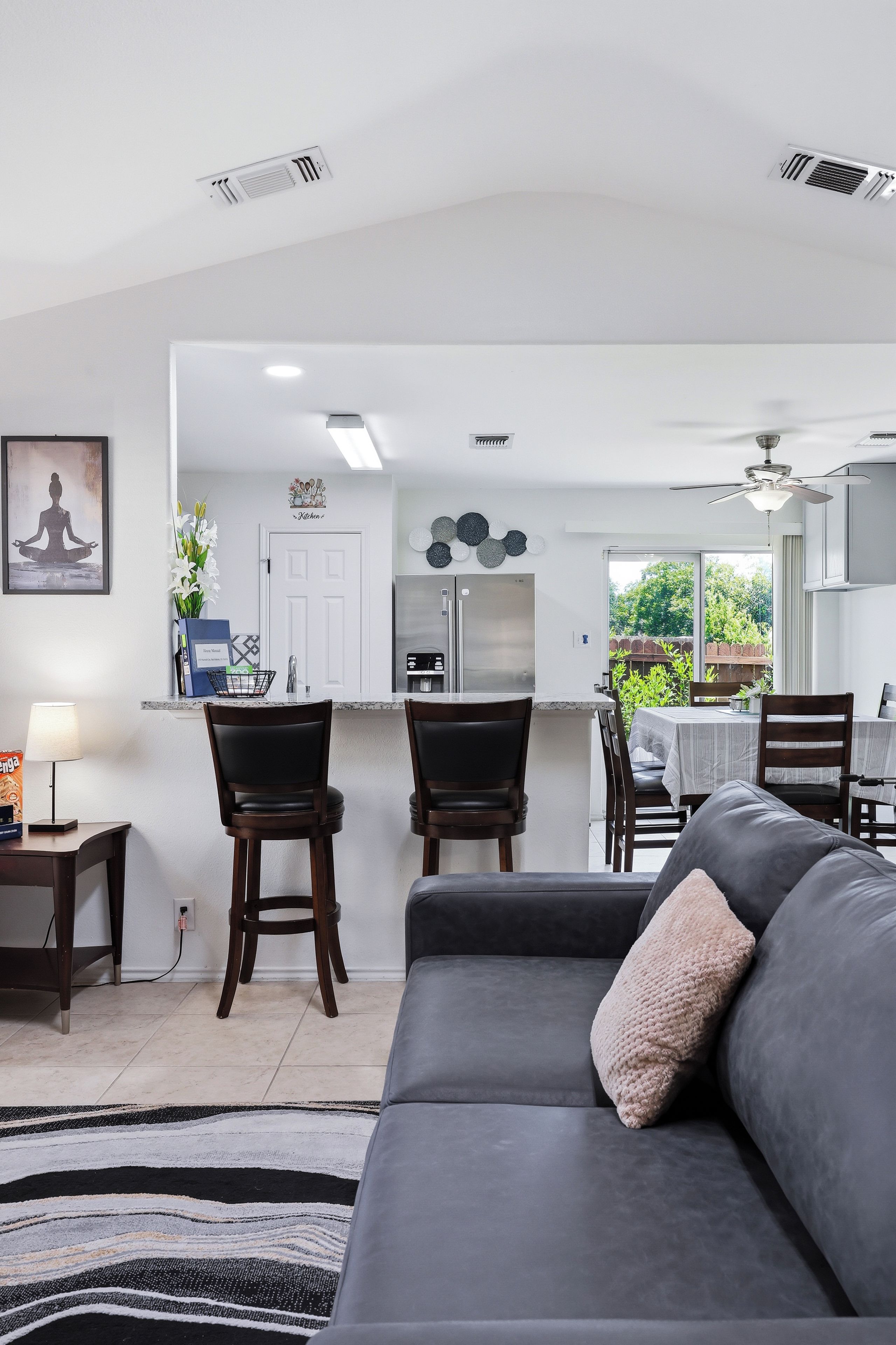 A modern living room with a grey sofa, a dining area with a table and chairs, and a kitchen with a refrigerator and a stove.