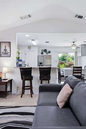 Un salon moderne avec un canapé gris, un coin repas avec une table et des chaises, et une cuisine équipée d’un réfrigérateur et d’une cuisinière.