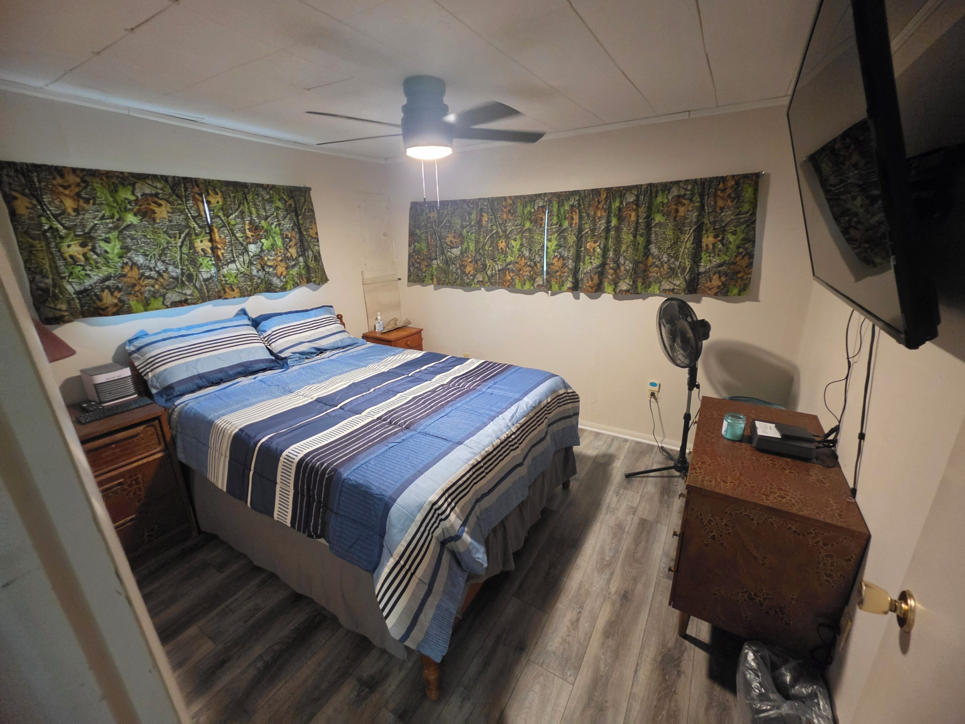 Un dormitorio con una cama, un escritorio, un ventilador y una televisión en la pared.