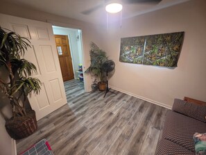 Un pasillo con piso de madera, una planta en maceta, un ventilador y un cuadro colgado en la pared.