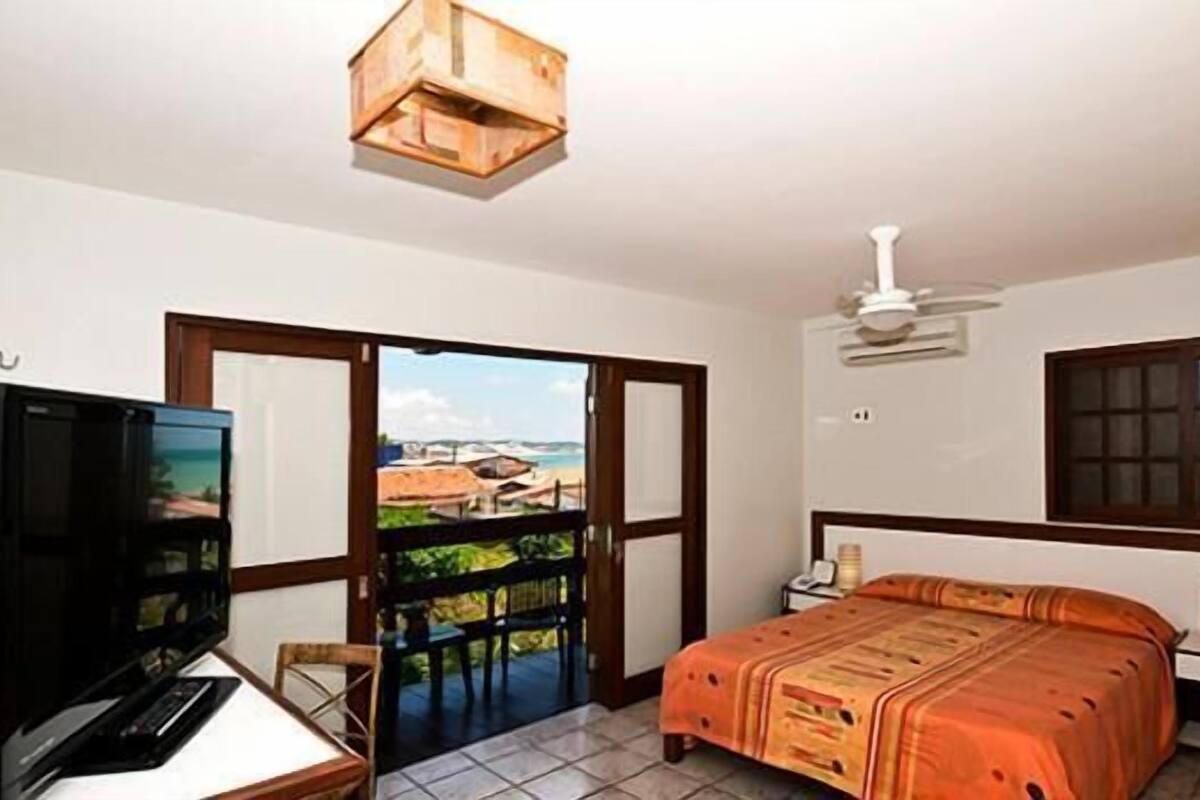 A hotel room with a bed, a desk with a computer, a television, a ceiling fan, and a window with a view of the beach.