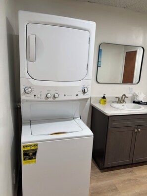 Un appareil compact combinant lave-linge et sèche-linge, doté d’un panneau de commande, d’un miroir au-dessus de l’évier et d’un meuble en dessous.