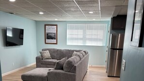 Un salon moderne avec un canapé d’angle, un téléviseur à écran plat, un tableau encadré au mur et un réfrigérateur.