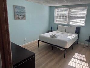 Een slaapkamer met een bed, een nachtkastje, een raam met jaloezieën en een schilderij aan de muur.