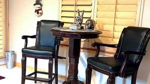 Een kamer met twee donkere houten stoelen met leren zittingen, een klein rond tafeltje met decoratieve objecten, en een raam met houten luiken.
