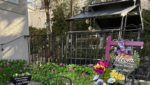 Een terras met een metalen tafel en stoelen, een decoratieve plantenbak met bloemen, en een bord met de tekst 'Welcome Please Watch Your Step!'