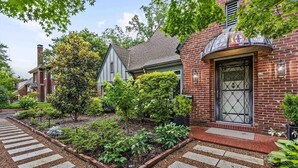 A brick house with a tiled entrance, a garden with a stone pathway, and a neighboring house with a gabled roof.