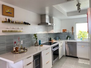 Eine moderne Küche mit weißen Schränken, einem Edelstahl-Geschirrspüler und einem eingebauten Backofen. Ein Fenster bietet Blick auf Grünflächen.