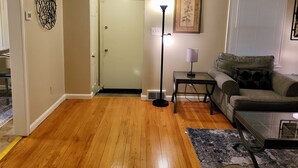 Un salon avec un plancher en bois, un canapé, une table basse, une lampe et une porte qui donne sur une autre pièce.