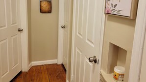 Un couloir avec un plancher en bois, des portes blanches et un paillasson noir. On y trouve un luminaire fixé au mur et une citation encadrée accrochée au mur.