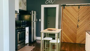 A compact kitchen with a dining area, featuring a wooden herringbone floor, a stainless steel refrigerator, and a 'Eat' sign above the door. - The Baum Suite: Charming 1950s Gem in Schulenburg ~New Owner~ (Schulenburg)