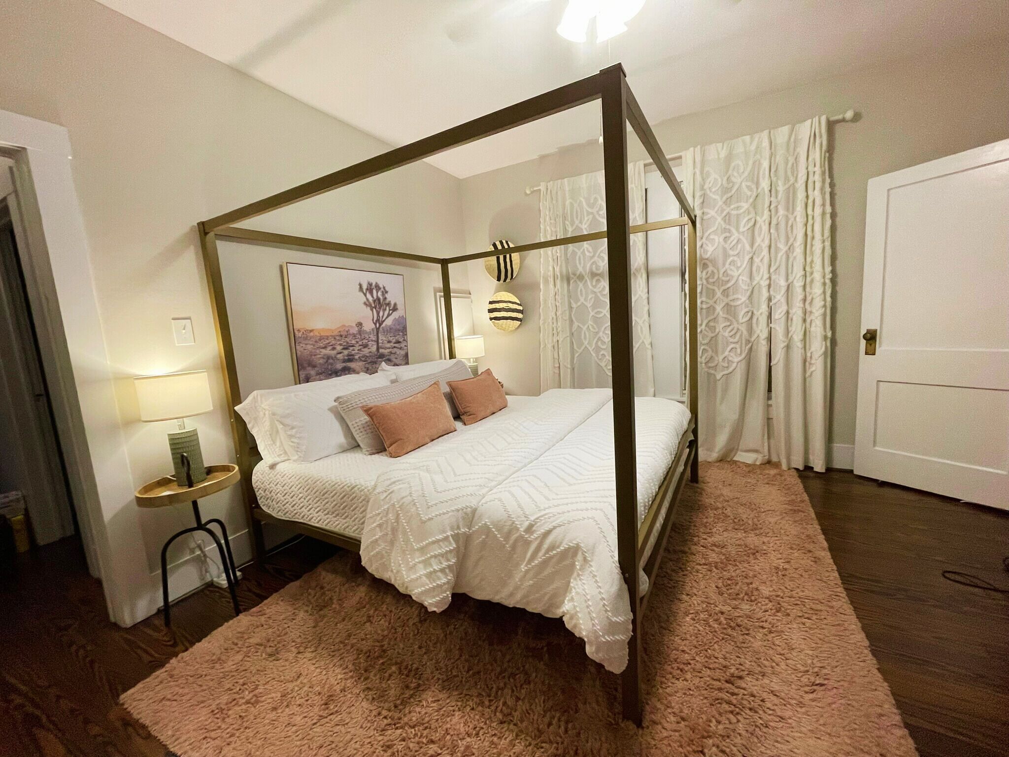 A four-poster bed with a white bedspread and two pillows, a bedside table with a lamp, and a framed picture on the wall.