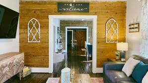 A modern living room with wooden panel walls, a central doorway, a blue sofa, a wooden coffee table, and a decorative sign above the doorway. - The Baum Suite: Charming 1950s Gem in Schulenburg ~New Owner~ (Schulenburg)