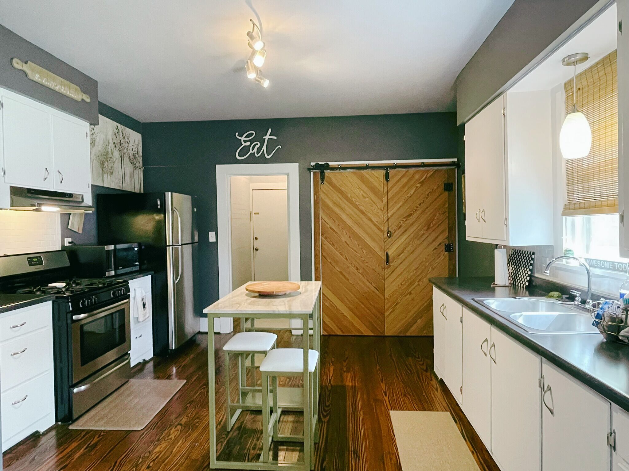 Une cuisine moderne avec des meubles blancs, une porte en bois à motifs en chevrons, un coin repas avec des tabourets et un plan de travail avec un évier.