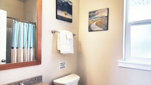 Een badkamer met een wastafel, toilet en een raam. Er hangen twee schilderijen aan de muur en een bordje met de tekst 'spoelen'.