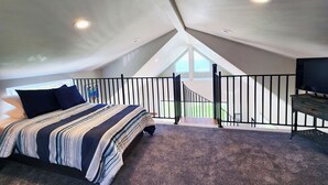 Een slaapkamer met een schuine kap, een bed met gestreepte beddengoed, een televisie en een balkon met een hekwerk.