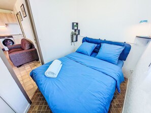 Un dormitorio pequeño y ordenado con una colcha azul, almohadas y una toalla enrollada. Se ve una silla y una lavadora en la habitación contigua.