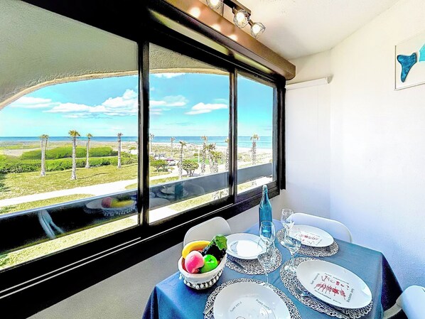 A dining table with plates, glasses, and a bowl of fruit, set against a view of a beach and palm trees.