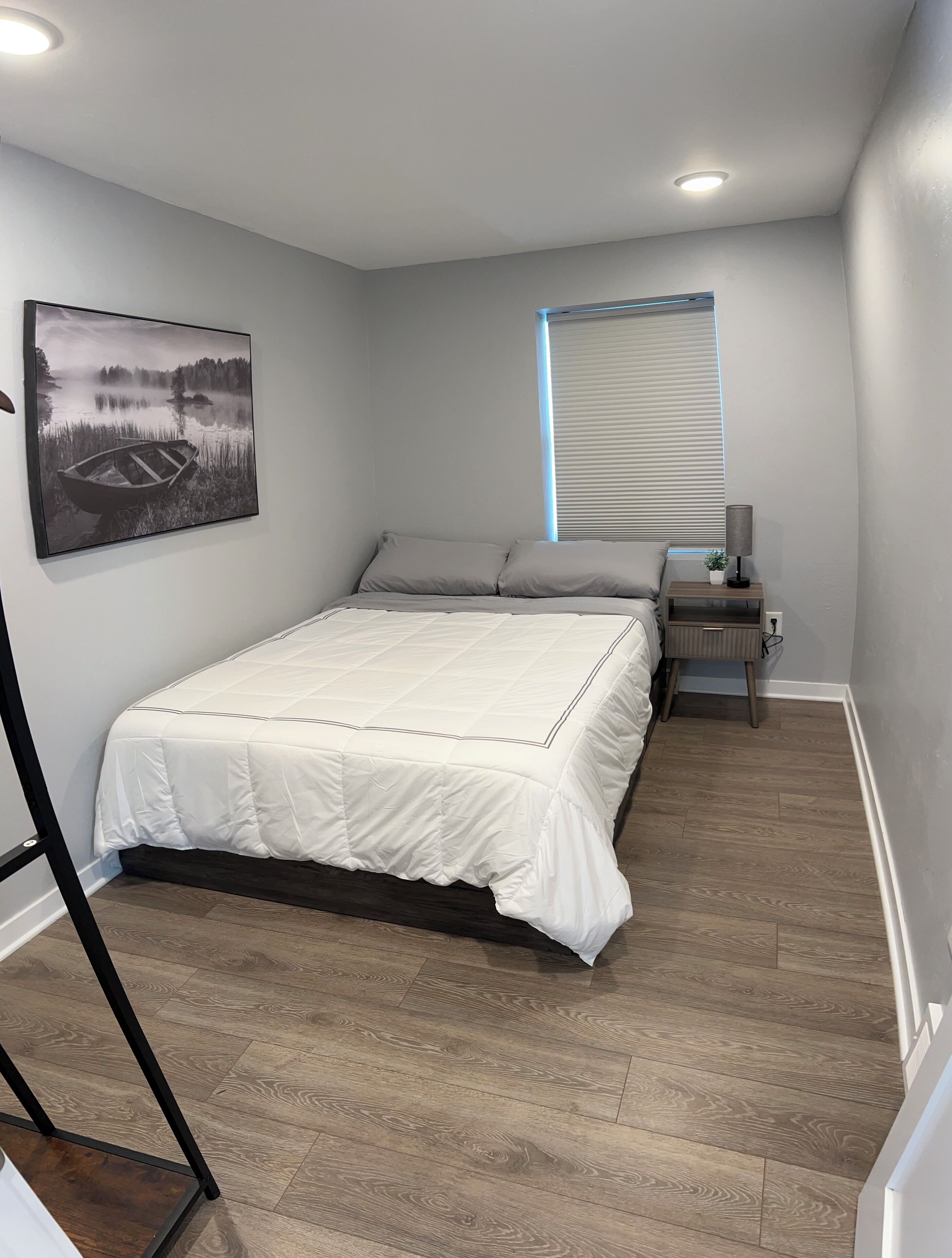 Ein Schlafzimmer mit Bett, Nachttisch, Lampe, Fenster mit Jalousien und einem gerahmten Bild an der Wand.