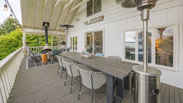 A covered outdoor deck with a long table and chairs, a grill, and large windows overlooking a body of water. - Beach Cottage w/ Water Access (Port Ludlow)
