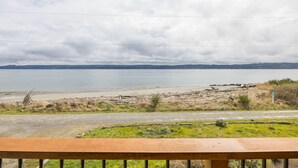 A view from a balcony overlooking a beach with a road leading to the water, a wooden fence, and a distant shoreline. - Waterfront Home w/ Beach access! (Port Ludlow)
