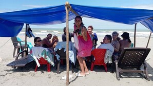 Un grupo de personas sentadas bajo un toldo azul en una playa arenosa, con vista al océano.