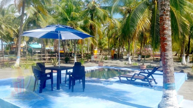 Zona de piscina con sillas de descanso y una mesa bajo una sombrilla azul, rodeada de palmeras.