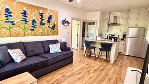 Een moderne woonkamer met een donkergrijze bank, twee bijpassende fauteuils en een houten salontafel. Achtergrond toont een keuken met witte kastjes en roestvrijstalen apparatuur.