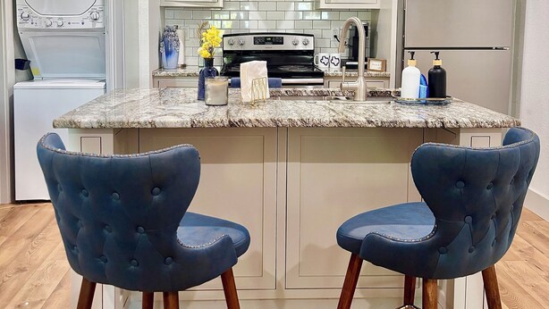 Een keuken met een centraal eiland, twee blauwe barstoelen, een roestvrijstalen oven en witte keukenkastjes.
