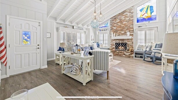 A spacious living room with a high ceiling, white walls, and a brick fireplace. There is a dining area with a white table and chairs, and a large window with a view of the ocean.